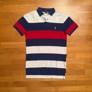 Randig pikétröja från Ralph Lauren - Säljer en snygg randig pikétröja från Ralph Lauren i blått, vitt och rött. Tröjan har en klassisk krage och korta ärmar. Den är prydd med det ikoniska broderade logotypen i gult på bröstet. Perfekt för en stilren look! Lite blekad vid kragen annars bra skick. Skick 8/10