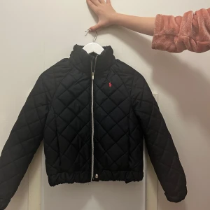 Svart quiltad jacka från Ralph Lauren - Säljer en snygg svart quiltad jacka från Ralph Lauren. Jackan har en dragkedja framtill och en liten logotyp i rött på bröstet. Perfekt för kyligare dagar med sin stilrena design och bekväma passform.