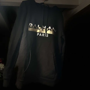 Svart hoodie från Balmain - Snygg svart hoodie från Balmain med guldtryck på bröstet. Den har en klassisk design med känguruficka och huva. Perfekt för en stilren look.