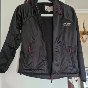 Svart jacka från On The Peak - Säljer en svart jacka från On The Peak med dragkedja och huva. Jackan har långa ärmar och praktiska fickor med dragkedjor. Perfekt för utomhusaktiviteter med sin sportiga design och funktionella detaljer.