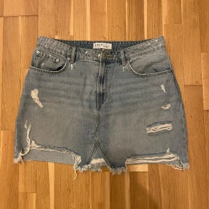 Blå jeanskjol från Primark - Säljer en cool blå jeanskjol från Primark med slitna detaljer och fransig nederkant. Kjolen har en klassisk femficksdesign och är perfekt för en avslappnad stil. Passar bra till sneakers eller sandaler för en trendig look.