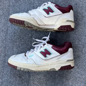 New Balance 550 burgundy blue  - Snygga New Balance 550 sneakers i vitt med vinröda detaljer. Skorna har en klassisk design med snörning och perforerade sidor för extra ventilation. Perfekta för en stilren och sportig look.