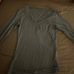 Grå långärmad topp från Intimissimi - Säljer en elegant grå långärmad topp från Intimissimi med vacker spetsdetalj vid halsringningen. Perfekt för en stilren look. Toppen är mjuk och bekväm, idealisk för både vardag och speciella tillfällen.