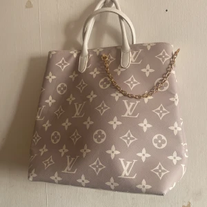 Beige handväska med monogram - Snygg beige handväska med vitt monogrammönster och guldkedja. Väskan har vita handtag och en praktisk dragkedja inuti. Perfekt för att ge din outfit en lyxig touch.