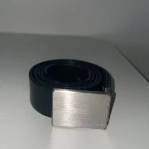 Svart bälte från Calvin Klein - Snyggt svart bälte från Calvin Klein med en stilren silverfärgad spänne. Perfekt för att ge en elegant touch till din outfit. Bältet är tillverkat i skinn och har en klassisk design som passar till många olika stilar.