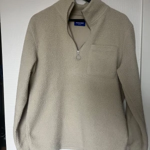 Beige fleece tröja från Jack & Jones - Säljer en mysig beige fleece tröja från Jack & Jones. Tröjan har en praktisk dragkedja framtill och en bröstficka. Perfekt för kyliga dagar.
