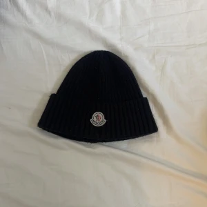 Svart ribbstickad mössa från Moncler - Svart ribbstickad mössa från Moncler med klassisk uppvikt kant och broderad logotyp framtill. Tillverkad i mjuk ull som håller dig varm och snygg under kalla dagar. Perfekt accessoar för en clean och stilren look.