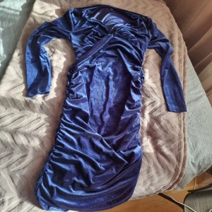 Blå sammetsklänning med rynk - Supersnygg långärmad sammetsklänning i djupblå färg med v-ringning och rynkade detaljer längs sidorna. Klänningen har en figurnära passform och känns lyxig tack vare det glansiga sammetstyget. Perfekt för dig som vill sticka ut med en elegant look.