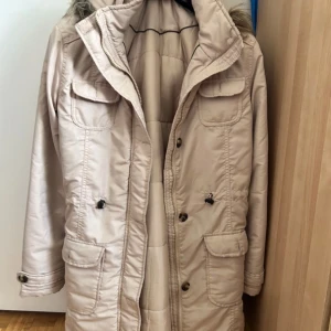 Beige parkasjacka med fuskpäls - Snygg beige parkasjacka med huva och fuskpälsdetalj. Jackan har flera fickor med lock och knappar, dragkedja framtill och justerbar midja med snörning. Perfekt för kyliga dagar och har en klassisk, stilren look.