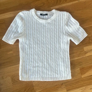 Vit kabelstickad tröja Gina Tricot - Vit kabelstickad tröja från Gina Tricot med korta ärmar och rund halsringning. Tröjan har ett klassiskt flätmönster och är gjord i mjukt stickat material. Perfekt för lager-på-lager och en clean look.