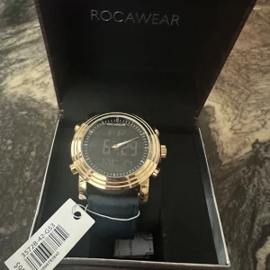 Guldig Rocawear klocka med blått band - Snygg klocka från Rocawear med rund urtavla i svart och guldig boett. Klockan har digital display och analoga visare, samt ett stilrent blått armband. Kommer i exklusiv trälåda med märkets logga. Perfekt accessoar för dig som gillar att sticka ut.