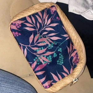 Beige och blå blommig necessär - Säljer en unik necessär med beige flätad utsida och blå tygpanel med rosa och gröna blommönster. Väskan har dragkedja, perfekt för småprylar eller smink. Superfin att ha i väskan eller som clutch till vardags.