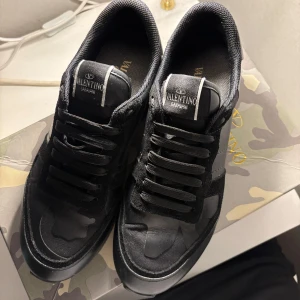 Svarta Valentino Garavani sneakers - Snygga svarta sneakers från Valentino Garavani med diskret camouflage-mönster. Skorna har snörning, rund tå och är tillverkade i en mix av skinn och textil. Perfekta för dig som gillar exklusiva och stilrena sneakers med en unik touch.