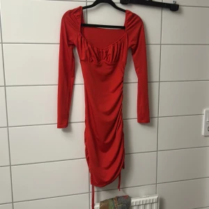 Röd långärmad klänning med rynk - Snygg röd klänning med långa ärmar och rynkade detaljer längs sidorna. Djup urringning och figurnära passform som framhäver siluetten. Perfekt för dig som vill sticka ut med en färgstark look.