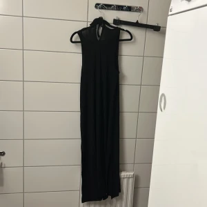 Svart långklänning med halterneck - En svart långklänning med halterneck och ärmlös design. Klänningen har en elegant och enkel siluett som faller snyggt längs kroppen. Perfekt för dig som gillar stilrena och tidlösa plagg.
