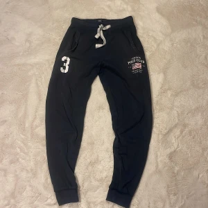 Blåa mjukisbyxor Vinson Polo Club S - Blåa mjukisbyxor från Vinson Polo Club med vita tryckdetaljer, snörning i midjan och muddar vid bensluten. Byxorna har fickor på sidorna och en sportig look med siffran 3 tryckt på ena benet. Perfekta för chill eller träning.