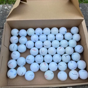 Titleist Mix - Bilden visar en kartong fylld med begagnade golfbollar av olika märken, främst Titleist. Perfekt för dig som vill öva din golf utan att spendera mycket pengar på nya bollar. Passar både nybörjare och erfarna golfare som vill ha extra bollar till träning eller spel. A/B klass, 50-pack.