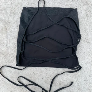 Svart glittrig snörad topp - Snygg svart topp med glittrigt tyg och långa snören som kan knytas kors och tvärs över ryggen. Toppen har bandeau-modell och är perfekt för dig som vill sticka ut med en trendig och edgy look. öppen i ryggen
