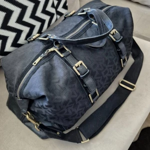 Svart resväska från DKNY med detaljer - Snygg svart resväska från DKNY med justerbar axelrem, guldfärgade spännen och dragkedja. Väskan har diskret mönster med DKNY-logga, förstärkta hörn i skinn och flera praktiska fack. Perfekt för weekendresor eller gymmet. ÄKTA 