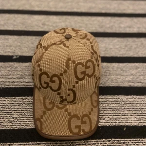 Beige keps från Gucci med GG-mönster - Snygg beige keps från Gucci med det klassiska GG-logomönstret i brunt över hela kepsen. Kepsen har böjd skärm och justerbar rem bak. Tillverkad i ett tåligt tygmaterial med mörkbrunt kantband runt skärmen för extra stilpoäng.