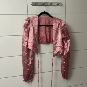 Rosa satin overshirt med knytband - Snygg croppad overshirt i glansigt rosa satinmaterial med långa ärmar och knytband framtill. Perfekt för att lyfta vilken outfit som helst med sin unika look och coola axelparti.