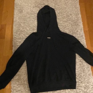 Svart hoodie från HUGO med logga - Navy blue hoodie från HUGO med stilren vit logga på bröstet. Klassisk modell med huva och dragsko, ribbade muddar och lång ärm. Perfekt för dig som gillar enkel och clean streetstyle. Mjuk bomullskänsla och snygg passform.