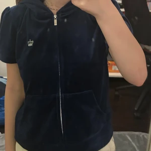 Mörkblå velourjacka Juicy Couture - Mörkblå velourjacka från Juicy Couture med korta ärmar, huva och dragkedja framtill. Klassisk kronbrodyr på bröstet och känguruficka. Perfekt för en chill och trendig look. Är en medium men passar som xs och xss.