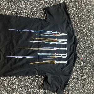 Svart AMIRI t-shirt med färgstänk - Svart t-shirt från AMIRI med coolt färgstänksmönster och stor logga på ryggen. Framsidan har en mindre version av samma tryck. Klassisk passform och rund hals. Perfekt för dig som vill sticka ut med en streetstyle-look.