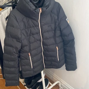 Svart pufferjacka från Svea - Säljer en svart pufferjacka från Svea med hög krage och snygga detaljer i guld på dragkedjorna. Jackan har broderad Svea-logga på bröstet och en patch med amerikansk flagga på ärmen. Perfekt för kalla dagar och riktigt skön att bära.