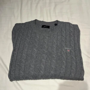 Grå stickad tröja från Gant - Snygg grå kabelstickad tröja från Gant med rund halsringning och diskret broderad logga på bröstet. Tröjan har klassisk passform och är perfekt för lager-på-lager-stil under kyliga dagar.