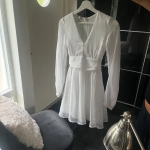 Vit omlottklänning från Bubbleroom - Supersöt vit kortklänning från Bubbleroom med omlott-look, v-ringning och långa ärmar. Klänningen har ett brett knytband i midjan och är gjord i ett lätt, flowy material som ger en romantisk vibe. Perfekt för sommarens alla tillfällen! Nypris är 699