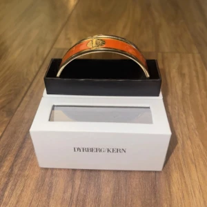 Orange armband från Dyrberg/Kern - Snyggt armband från Dyrberg/Kern i orange med guldfärgade detaljer och rund logga framtill. Armbandet är stelt och har en modern, stilren design som sticker ut på handleden. Perfekt accessoar för dig som vill addera färg till din look.