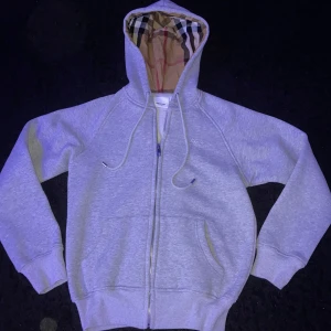 Grå hoodie från Burberry med dragkedja - Grå hoodie från Burberry med klassiskt rutigt foder i huvan och broderad logga på bröstet. Tröjan har dragkedja, snörning i huvan och två fickor framtill. Perfekt för dig som gillar stilren streetwear med lyxig touch! Tröjan är använd 1 gång.