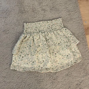 Blommig volangkjol - Supersöt kort kjol med blommigt mönster i ljusa färger och volanger. Kjolen har bred smock i midjan för en bekväm passform och är perfekt för varma dagar.