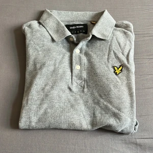 Grå pikétröja från Lyle & Scott - Snygg grå pikétröja från Lyle & Scott med klassisk krage och tre knappar framtill. Tröjan har det ikoniska gula fågel-loggan broderad på bröstet. Tillverkad i mjuk bomull som känns skön mot huden. Perfekt för en clean och stilren look. Hör av er vid minsta lilla fråga. 