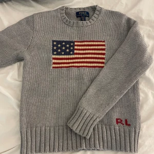 Grå Polo Ralph Lauren tröja - Jättesnygg grå stickad tröja från Polo Ralph Lauren med amerikansk flagga framtill och RL-broderi i rött vid nederkanten. Jättekänd och brukar vara slutsåld. Jag köpte den för 2000 kr. Ja säljer för 1000 men pris kan diskuteras. Använd som mest 5 gånger och jag säljer den för att den tyvärr blivit liten 