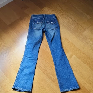 Blå bootcut jeans från H&M - Snygga blå bootcut jeans från H&M med låg midja och justerbar midja. Klassisk femficksmodell med knappar och dragkedja framtill. Jeansen har en lätt tvättad look och bakfickor med lock och knapp.