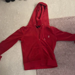 Röd Juicy Couture velour hoodie S - Röd velour hoodie från Juicy Couture med glittrande stenar på bröstet och stor Juicy-text i strass på ryggen. Dok är dragkedjan av och går inte att lösa
