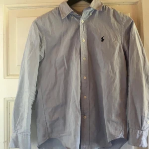 Blå skjorta från Ralph Lauren - Klassisk ljusblå skjorta från Ralph Lauren med broderad logga på bröstet. Skjortan har lång ärm, knappar framtill och är tillverkad i bomull. Perfekt för en clean och stilren look. Storlek 14 (44) ca 10-14 år