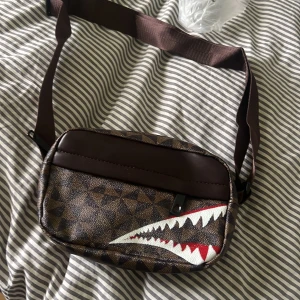 Brun axelväska med hajmotiv - Säljer en brun axelväska med geometriskt mönster och ett coolt hajtandsmotiv i vitt och rött på framsidan. Väskan har justerbart band och dragkedja upptill. Perfekt för att bära det viktigaste och ge outfiten en edgy vibe. Det finns en svart också.