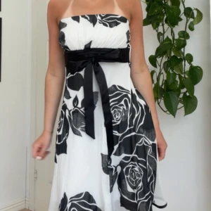 Vit och svart axelbandslös klänning - Snygg axelbandslös klänning i vitt med stora svarta rosor och ett brett satinband i midjan som knyts framtill. Man kan även knyta banden bakom ryggen. Klänningen har en utsvängd kjol och slutar strax ovanför knäna. Står varken märke eller storlek men den sitter som en XS