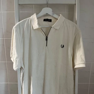 Fred Perry - Zip upp T-shirt  - Snygg vit pikétröja från Fred Perry i frotté med klassisk krage och half zip-dragkedja framtill. Tröjan har korta ärmar och den ikoniska Fred Perry-loggan broderad på bröstet. Perfekt för en clean och sportig stil.