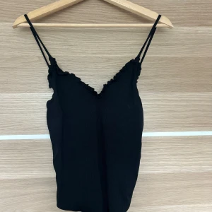 Svart ribbad body med volang - Svart ribbad linnetopp med smala axelband och söt volangkant längs v-ringningen. Toppens material känns mjukt och stretchigt, perfekt för en stilren look. Enkel att matcha med jeans eller kjol för en trendig outfit. 