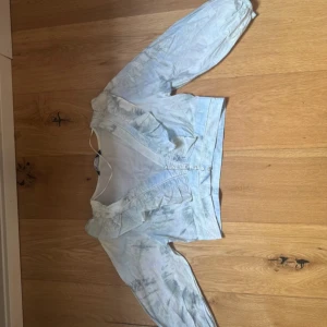 Ljusblå blus med volang och knappar - Supersöt ljusblå blus med volangdetaljer längs v-ringningen och små knappar framtill. Blusen har långa ärmar med resår vid handlederna och är gjord i ett tunt, lätt tyg. Perfekt för dig som gillar romantisk och bohemisk stil.