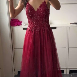 Vinröd balklänning med spetsdetaljer - Vinröd långklänning med smala axelband och v-ringning. Överdelen har vackra spets- och broderidetaljer med lite glitter, och kjolen är i flera lager tunn tyll som ger en drömmig känsla. Perfekt för bal eller student. Sitter snyggt och har en elegant siluett.