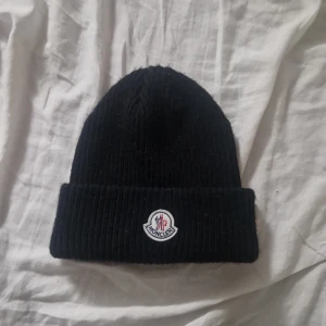 Svart Moncler ribbstickad mössa - Svart ribbstickad mössa från Moncler med klassisk uppvikt kant och broderad logotyp framtill. Perfekt accessoar för kyliga dagar och ger en clean look till din outfit.