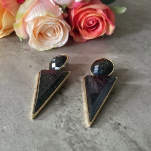 Svarta glittriga triangulära örhängen - Snygga örhängen i svart med glittrig finish och guldiga kanter. De har en cool geometrisk design med rund överdel och spetsig triangelformad nederdel. Perfekta för att ge din look en edgy och trendig touch.