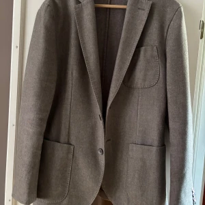 Grå  kavaj från Massimo Dutti - Stilren grå kavaj från Massimo Dutti i klassisk modell med tre utanpåfickor och snygga knappar vid ärmslutet. Tillverkad i ett mjukt ullmaterial som ger en exklusiv känsla. Perfekt för dig som vill ha en tidlös och clean look.