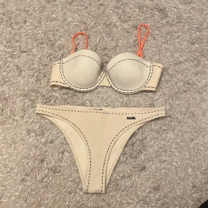  Triangl bikini - Superfin vit bikini från Triangl med ribbad struktur och svarta sömdetaljer. Överdelen har bygel och vadderade kupor samt orange axelband som ger en snygg kontrast. Underdelen har samma ribbade tyg. Perfekt för stranden eller poolen!