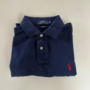 Marinblå pikétröja från Ralph Lauren - Klassisk marinblå pikétröja från Polo Ralph Lauren i bomull. Custom fit-modell med korta ärmar, krage och två knappar framtill. Ikoniska röda loggan broderad på bröstet. Perfekt för en clean och stilren look.
